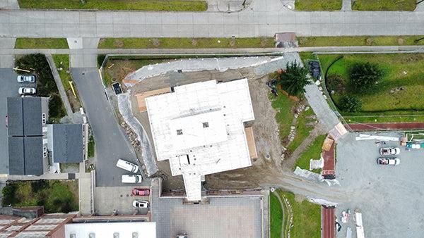 鸟瞰正在建设的建筑，显示与现有建筑的狭窄连接, a road at the top of the image, and a parking lot to the left